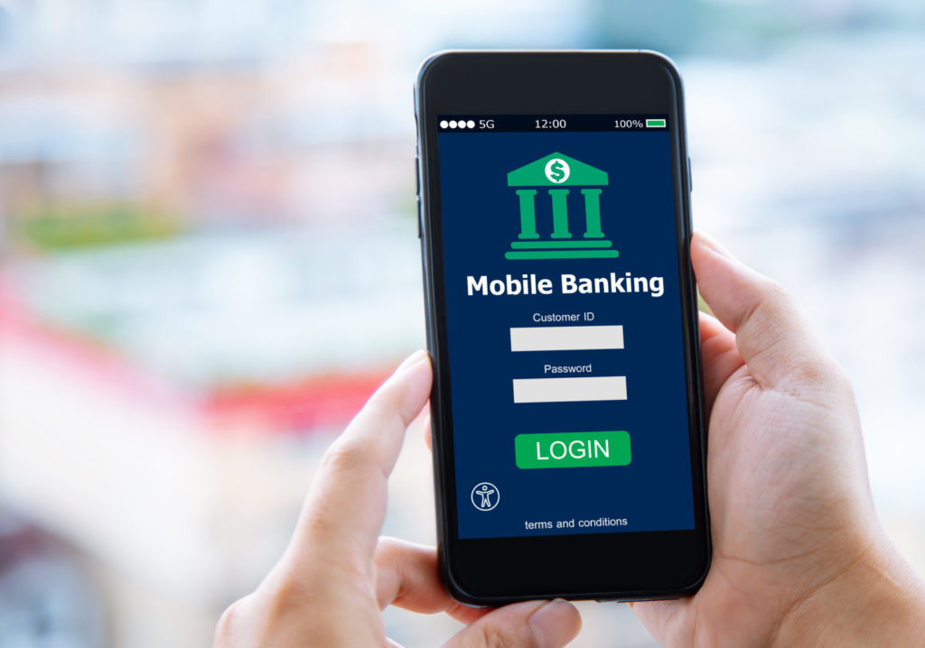 Hands holding a cell phone with a mobile banking login open, and an accessibility overlay icon lower left of screen.