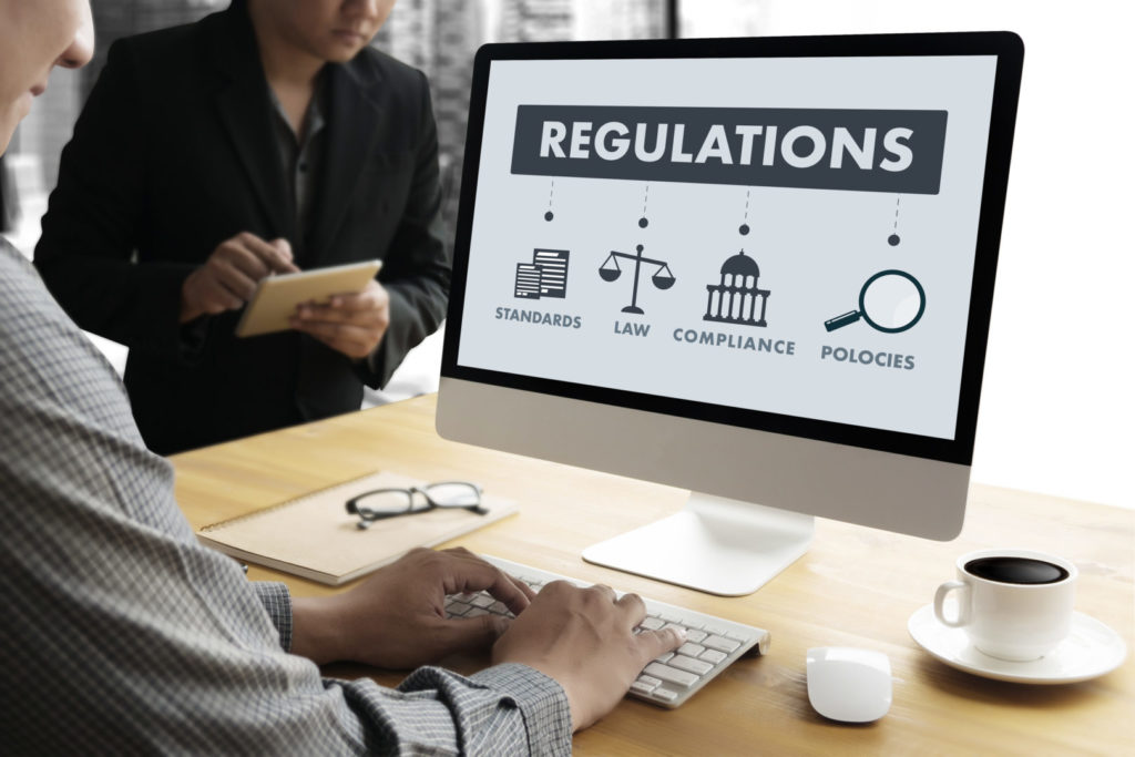 Two business people in front of a PC with the word REGULATIONS in big font.