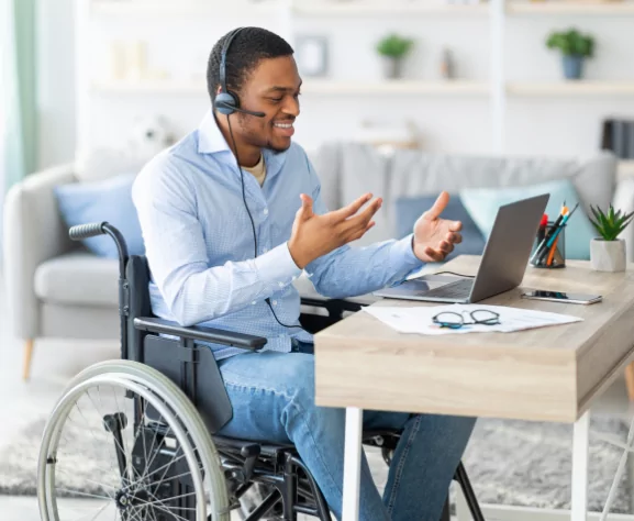 Black man with headset in a wheelchair, gesturing to a laptop.