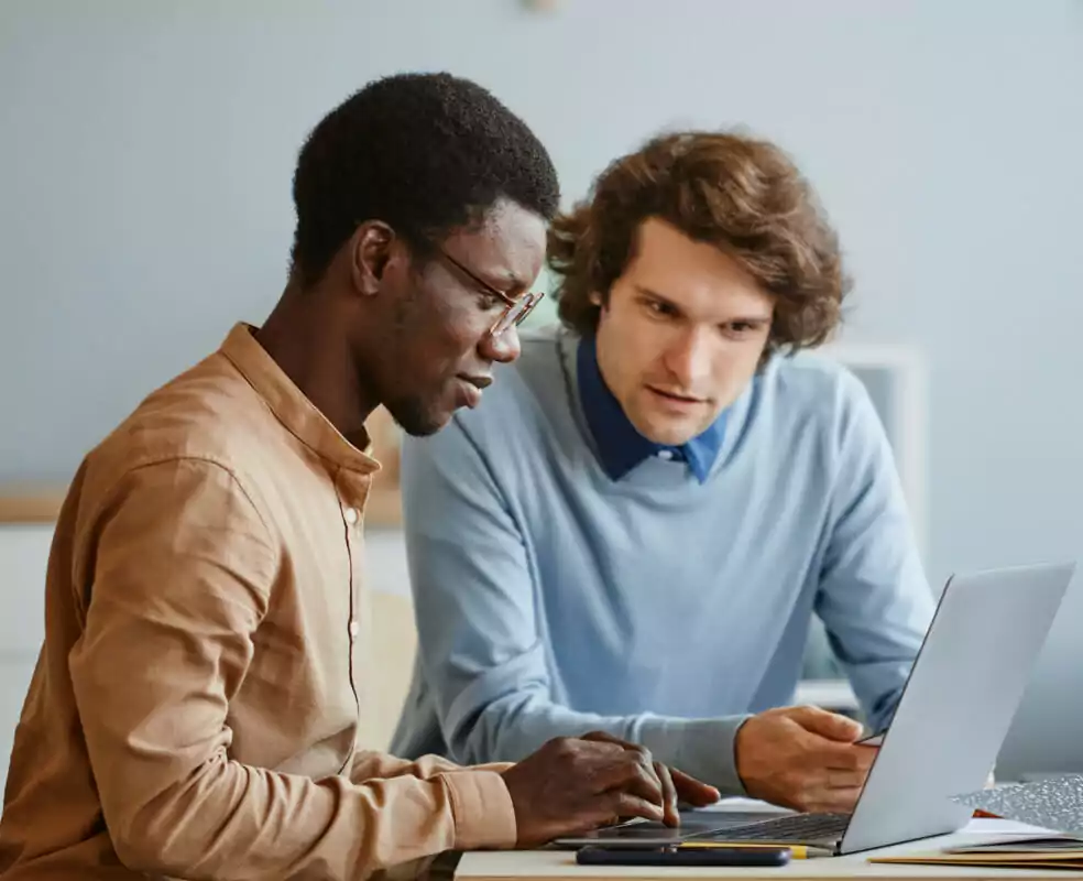 Two men sit at a desk in front of a computer. One wears a tan buttoned-up shirt, and the other wears a light blue sweater.