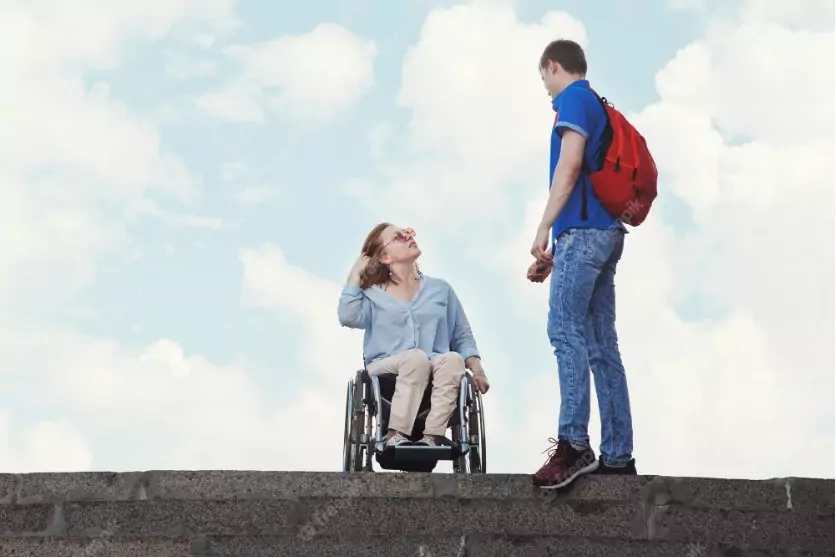 Blonde woman wearing glasses in a wheelchair looks up at a man wearing a backpack. Behind them is a blue sky with white clouds.