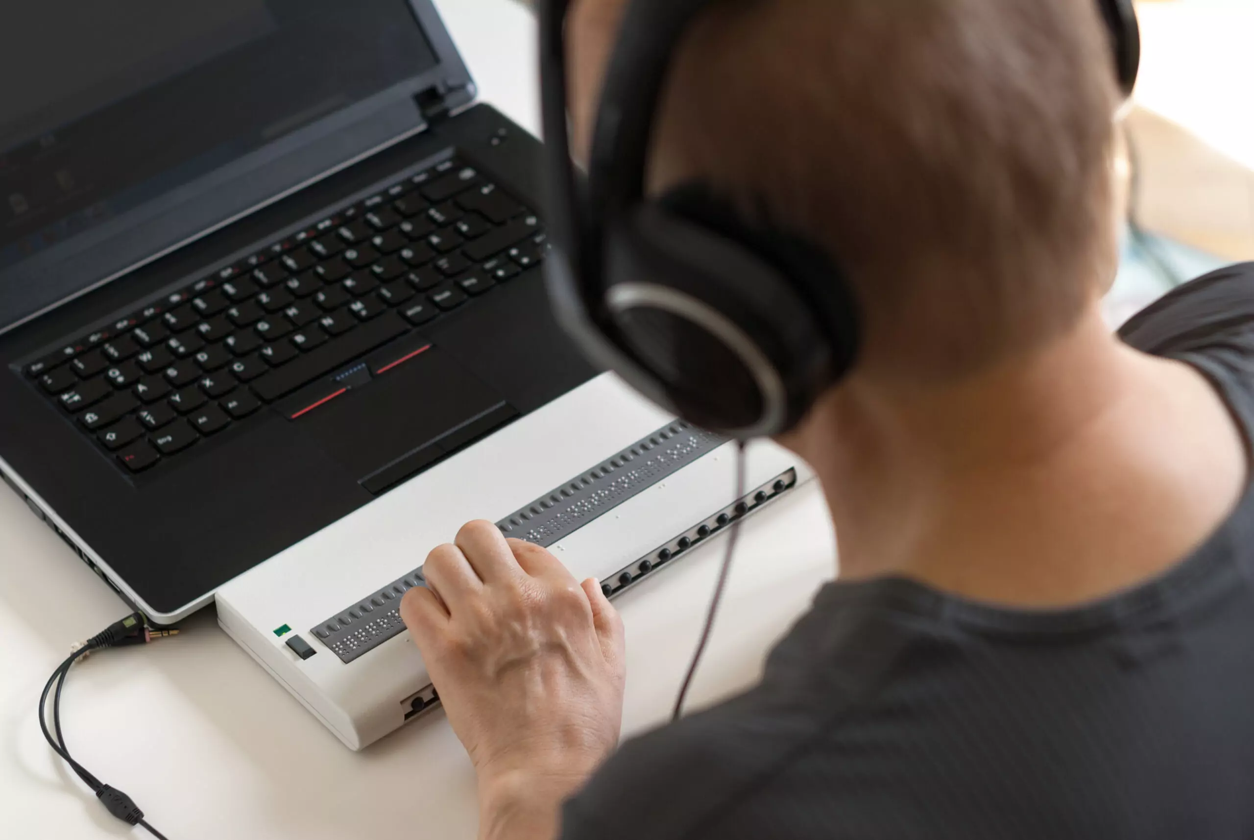 Looking over the shoulder of a man using a braille reader attached to a laptop. He is wearing headphones.