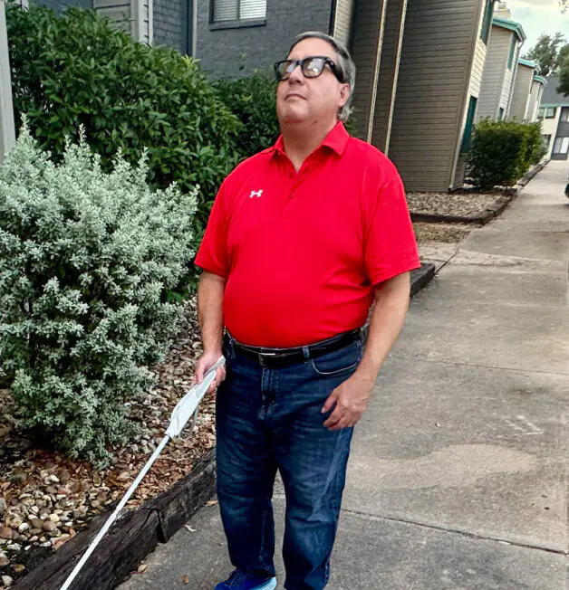 A man wearing black glasses and a red shirt is using a white cane while walking down a sidewalk.